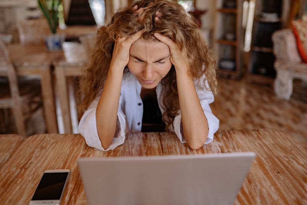 A stressed woman sits at a wooden table, frustrated with her laptop work.